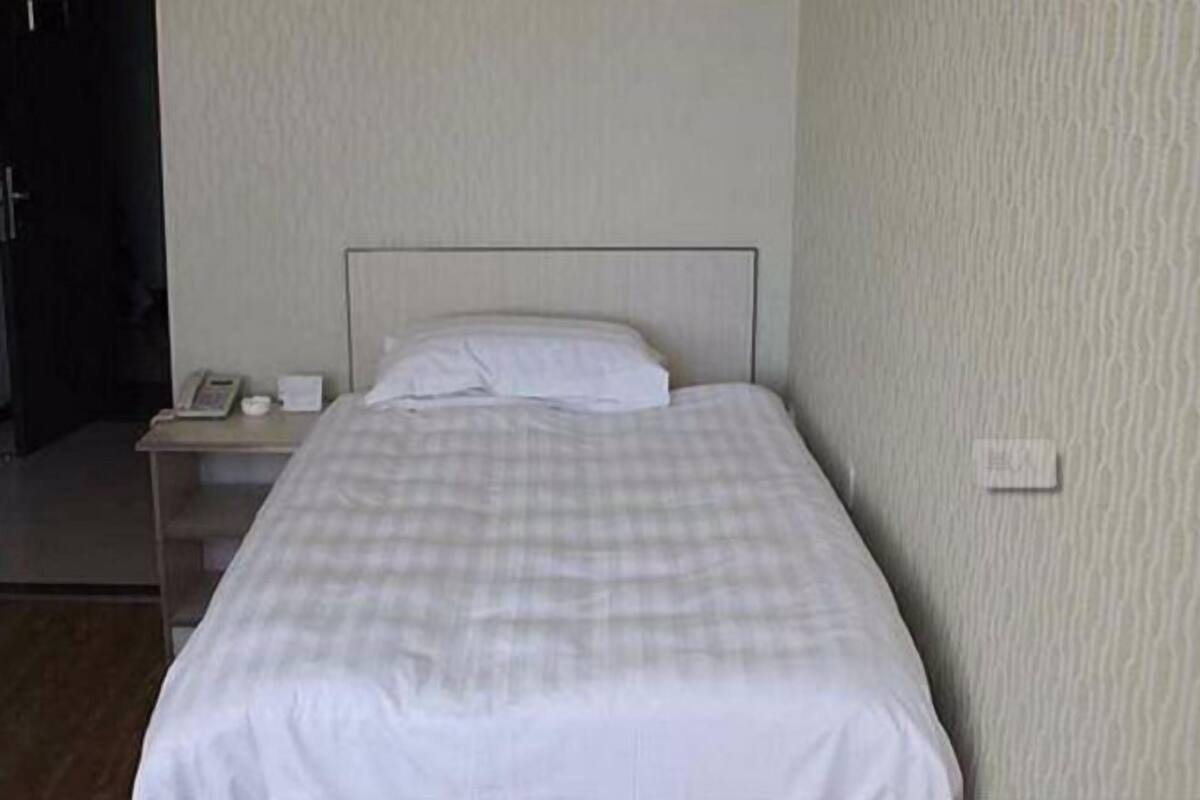 A single bed with a checkered bedspread, a bedside table with a phone, and a wall-mounted light switch.