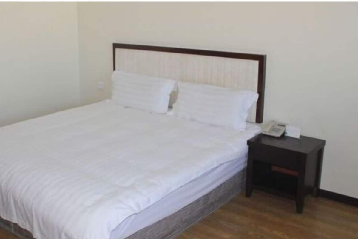 A single bed with white bedding, a wooden headboard, a small bedside table with a phone and papers, and a plain wall.