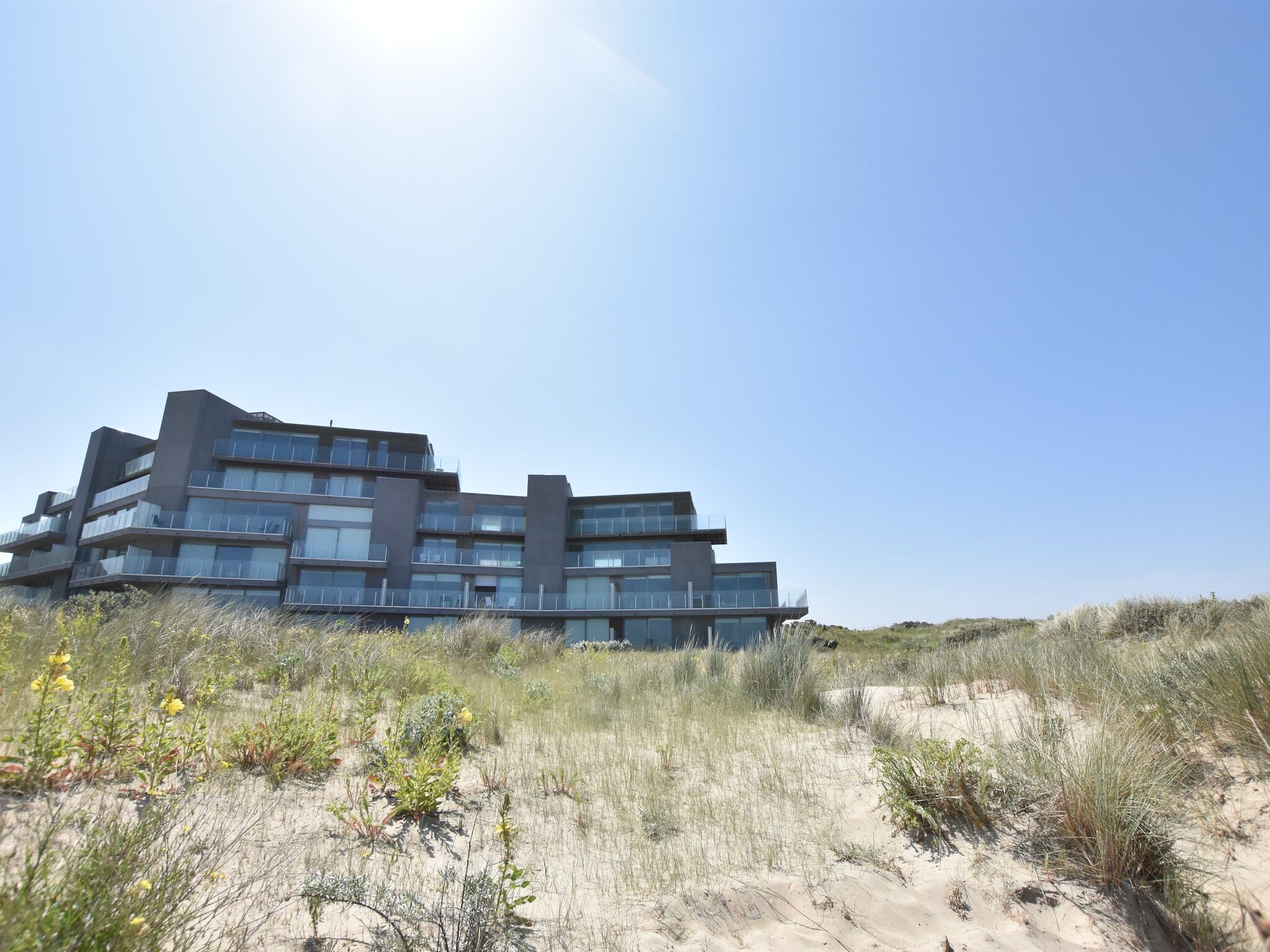 Un immeuble moderne à plusieurs étages, doté de balcons, situé sur une dune de sable où la végétation est clairsemée.