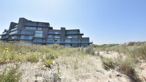 A modern multi-story building with balconies, situated on a sandy dune with sparse vegetation.