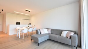 A modern living room with a grey sectional sofa, a dining area with white chairs, and a kitchen with white cabinets.
