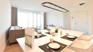 A modern dining area with a white table set for two, a sofa, and a large window with curtains.