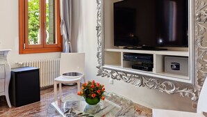 Een moderne woonkamer met een grote flatscreen televisie, een kleine koelkast, een witte stoel en een glazen salontafel met een bloemenvaas.