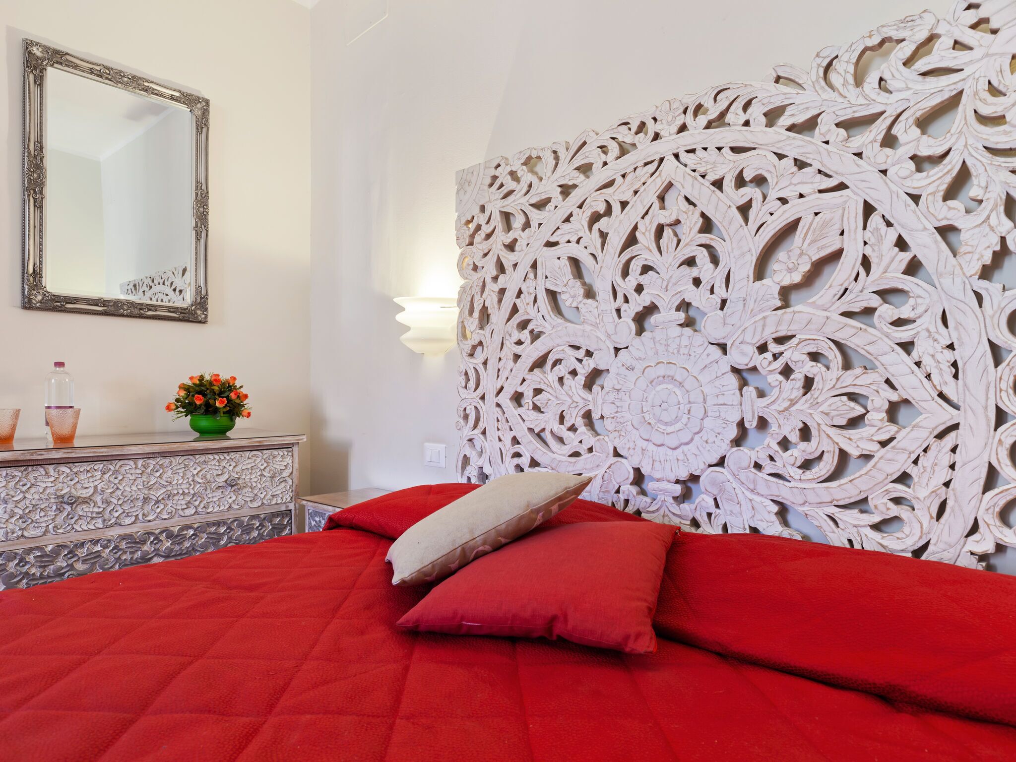 A bedroom with a red bedspread, a decorative headboard, a mirror, and a dresser with a vase of flowers.