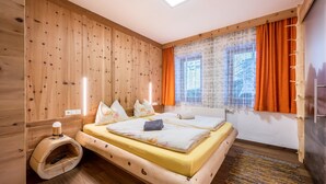 Ein Zimmer mit Holzwänden, einem Bett, Kissen, einem kleinen Nachttisch und einem Fenster mit Vorhängen.