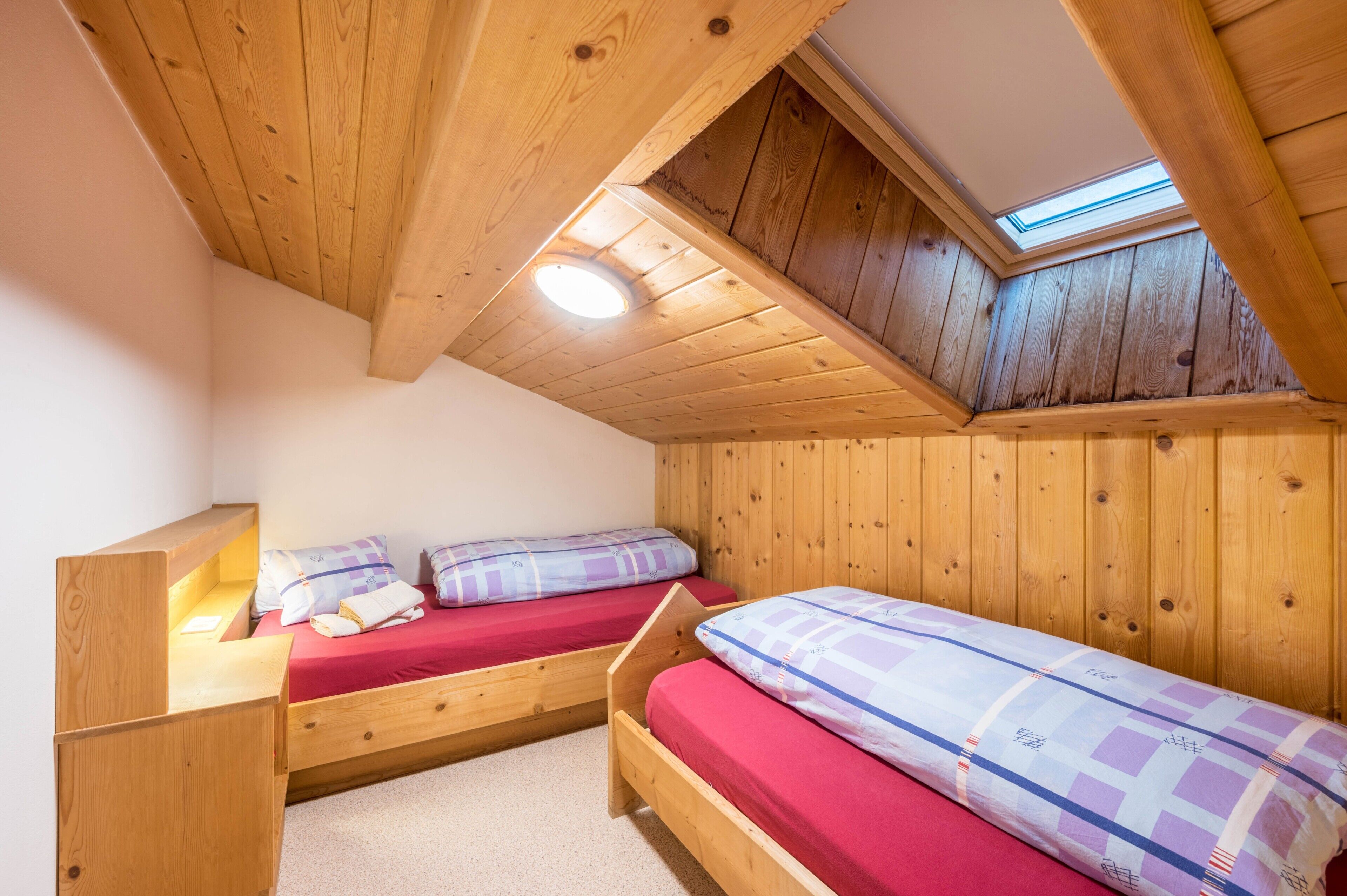 Ein kleiner Raum mit Holzboden und -wänden, zwei Betten, einem Oberlicht und einer geneigten Decke.
