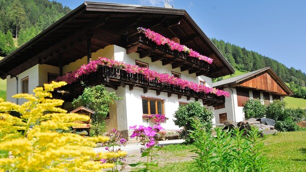 En traditionel bjælkehytte med balkoner pyntet med blomster, omgivet af frodig vegetation og et bjerglandskab i baggrunden.