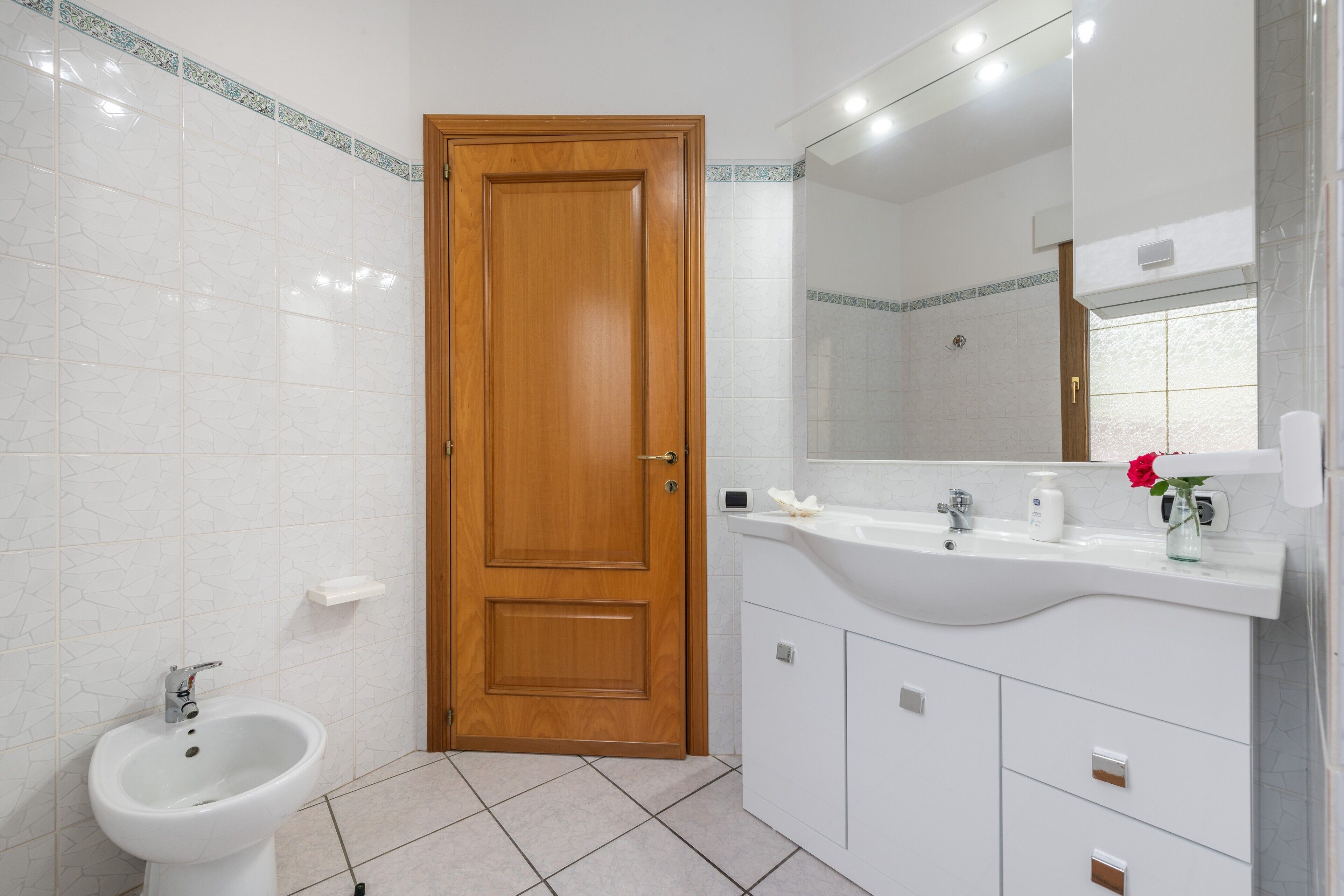 Une salle de bain avec une porte en bois, des carreaux blancs, un lavabo, un miroir et un bidet.