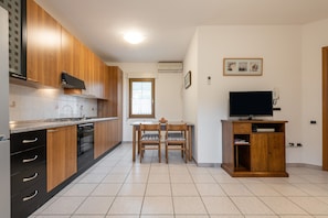 Une cuisine avec des meubles de rangement en bois, une table à manger avec des chaises, un téléviseur posé sur un meuble et une fenêtre.