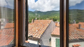 Vista dalla finestra su un paesaggio montuoso con tetti in terracotta e cielo sereno.