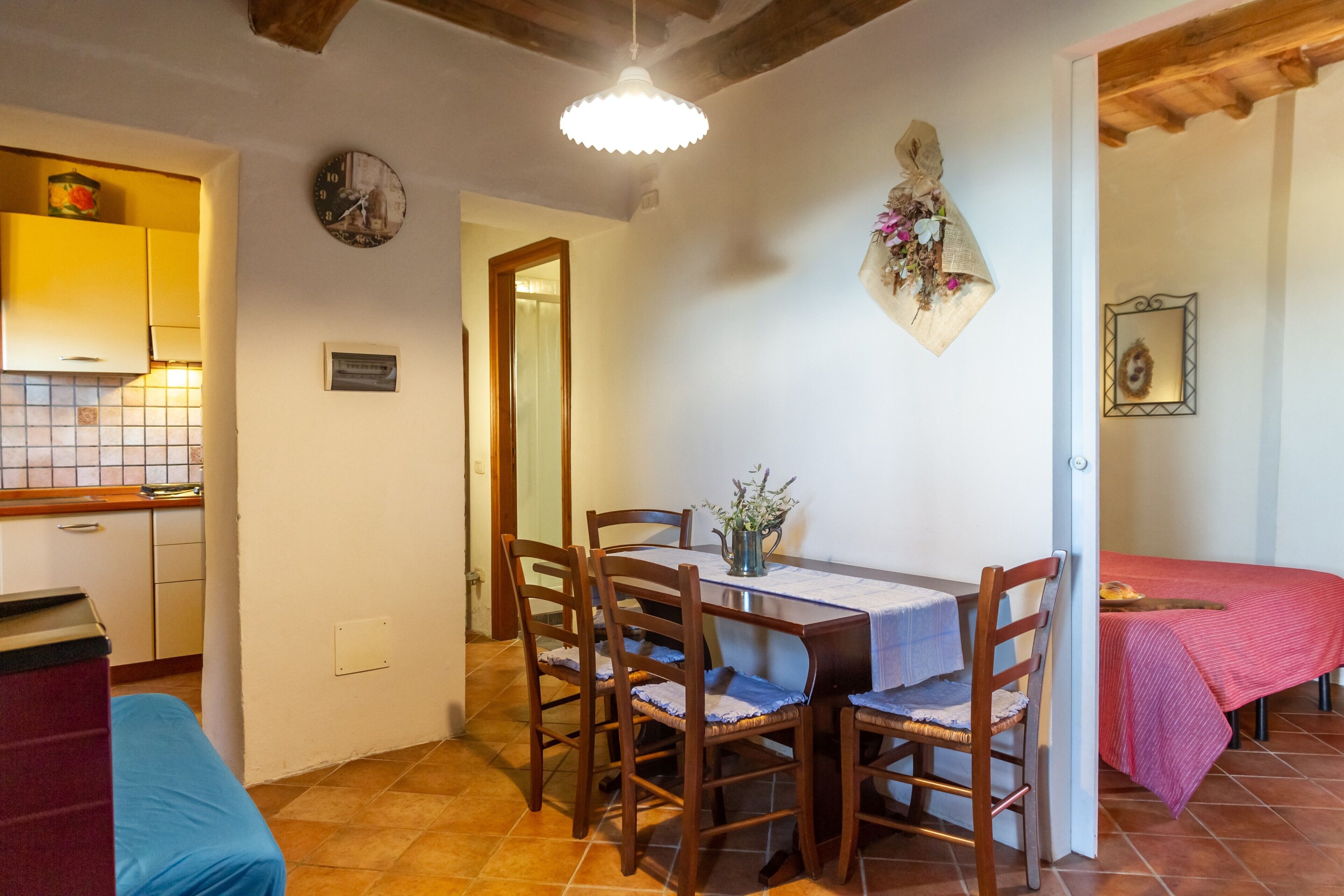 A dining area with a wooden table and chairs, a kitchen in the background, and a bed with a red blanket.