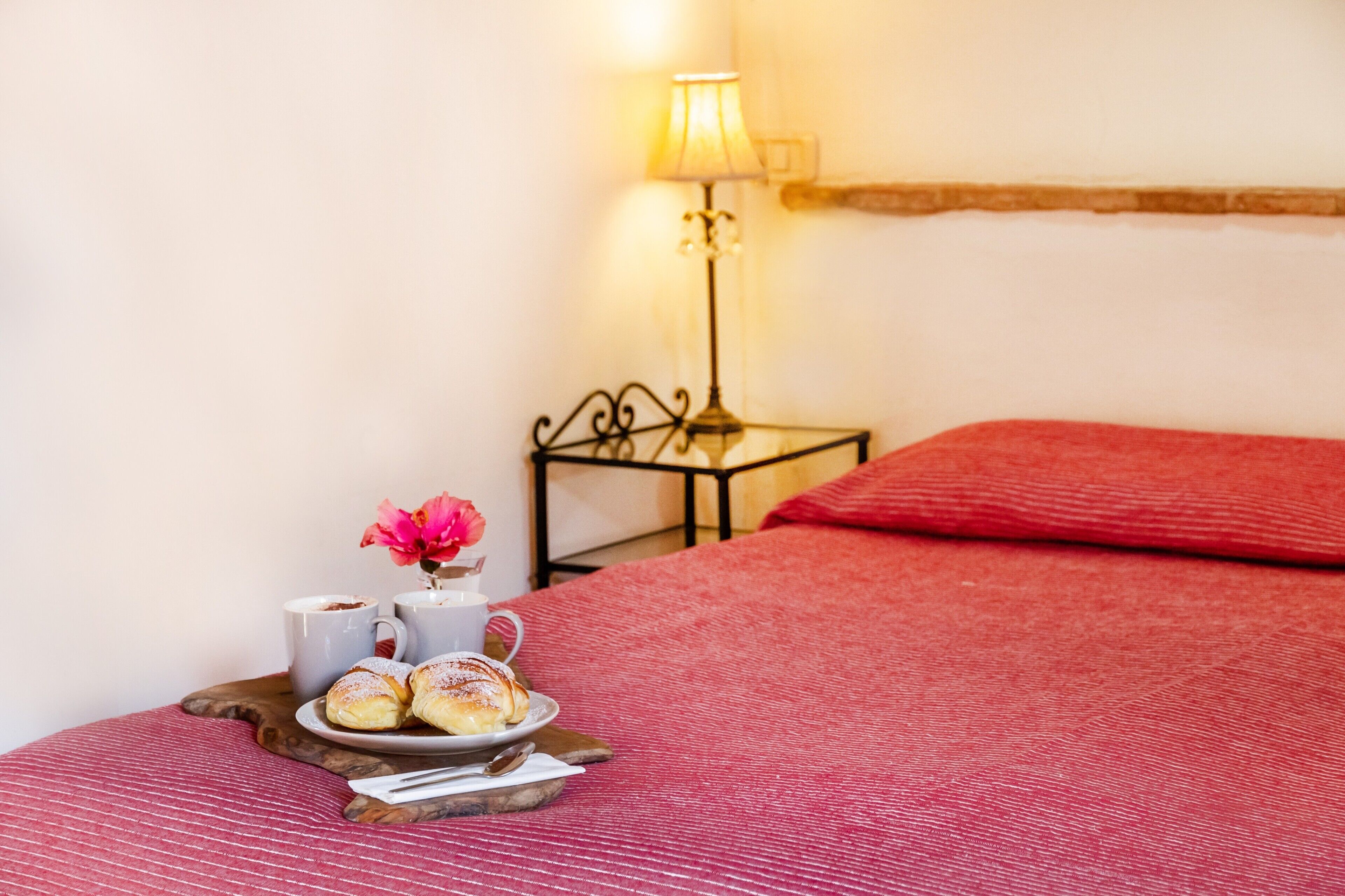 Un lit bien fait, recouvert d’une couverture rouge, une petite table de chevet avec une lampe, une assiette de pâtisseries et une tasse de café.