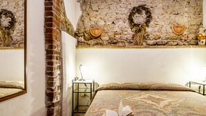 Een slaapkamer met een bedovertrek met patroon, een bakstenen muur, een stenen muur met decoratieve details, een nachtkastje met een lamp en een spiegel aan de muur.