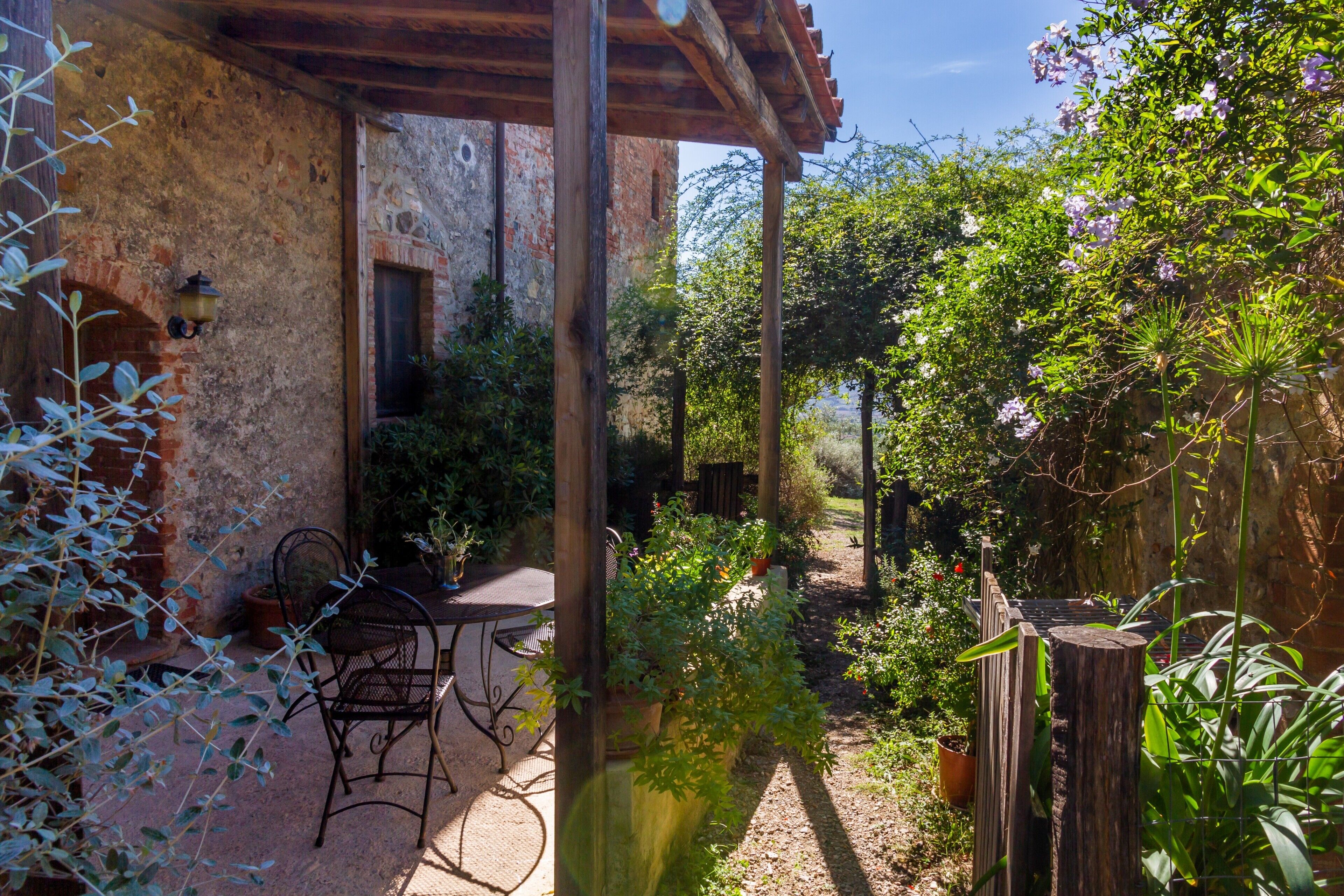 A rustic outdoor patio with a table and chairs, surrounded by greenery and a stone wall.