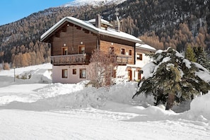 A wooden chalet with a balcony, surrounded by snow-covered ground and a forested mountain backdrop.