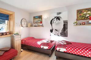 Une chambre avec deux lits, une commode en bois, une fenêtre donnant sur un beau paysage, et des éléments de décoration murale, notamment une horloge et un tableau représentant un chat.