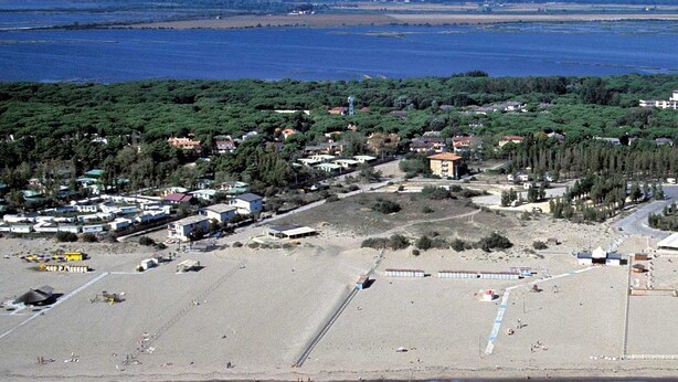 Vue aérienne d’une zone côtière avec une plage de sable, quelques bâtiments et une étendue d’eau en arrière-plan.