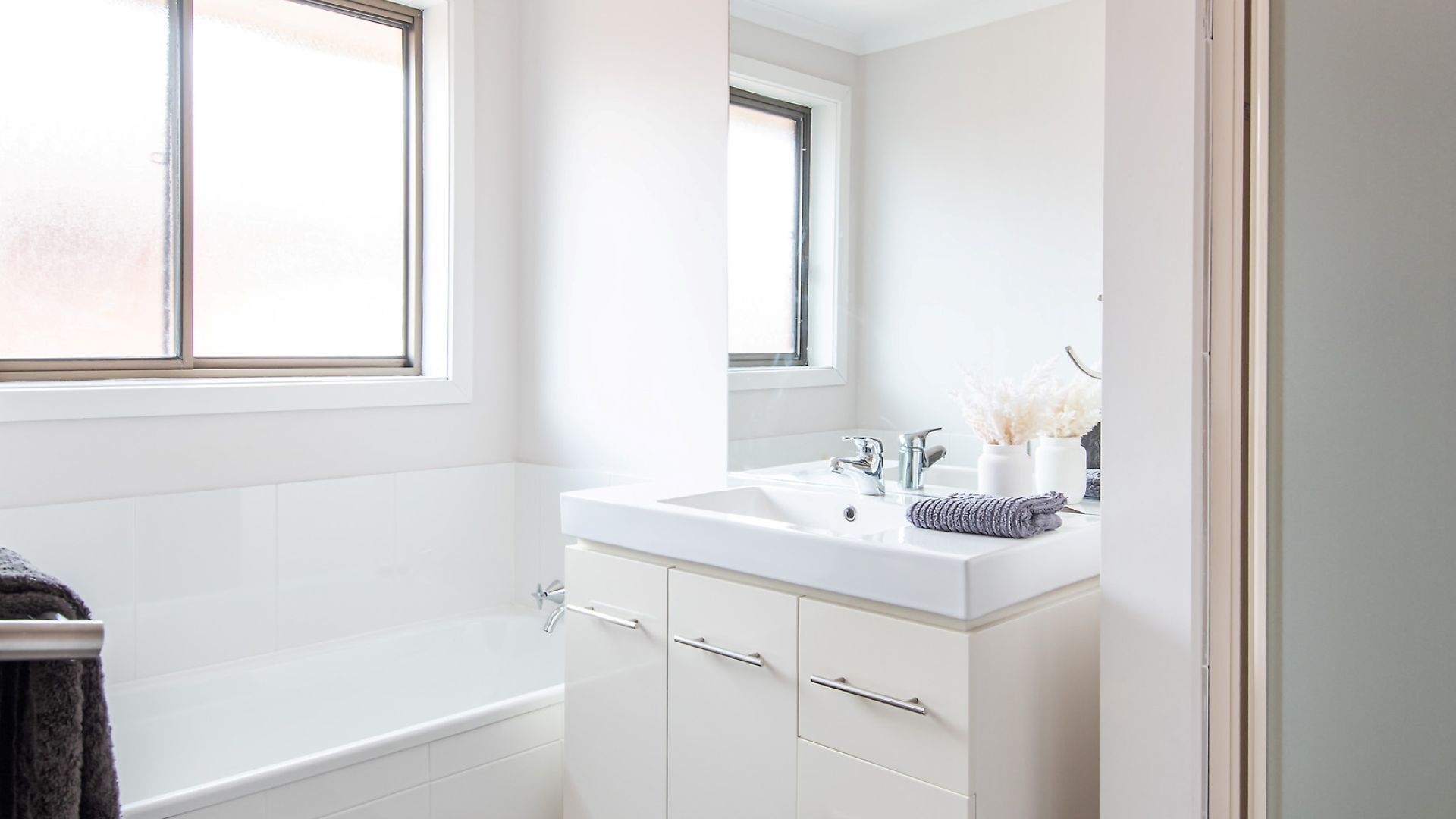 A modern bathroom with a white sink, mirror, and bathtub. There are two windows providing natural light.