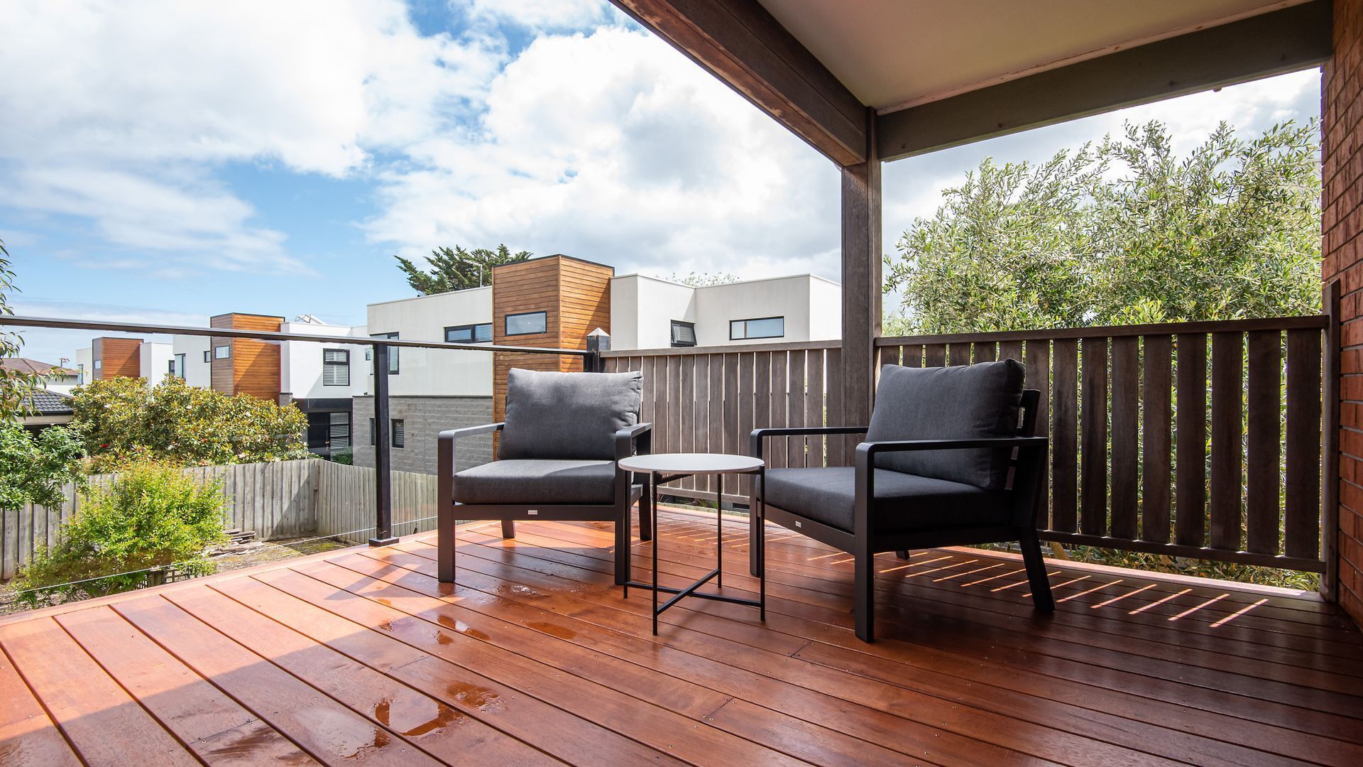 Une terrasse en bois avec deux chaises et une petite table, entourée d’une clôture en bois et offrant une vue sur les maisons voisines.