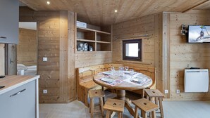 Interior acogedor de una cabaña de madera con una mesa redonda para comer rodeada de taburetes, una estantería empotrada con utensilios de cocina, una ventana pequeña y un televisor montado en la pared.