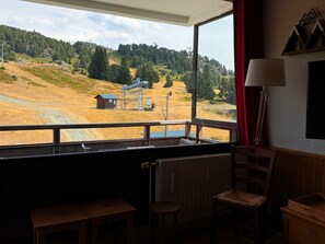 A room with a large window overlooking a rural landscape, a wooden chair, a small table, and a lamp.