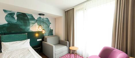 Una habitación de hotel con una cama, un sillón gris, un sillón rosa, una mesa redonda y una pared decorada con arte abstracto.