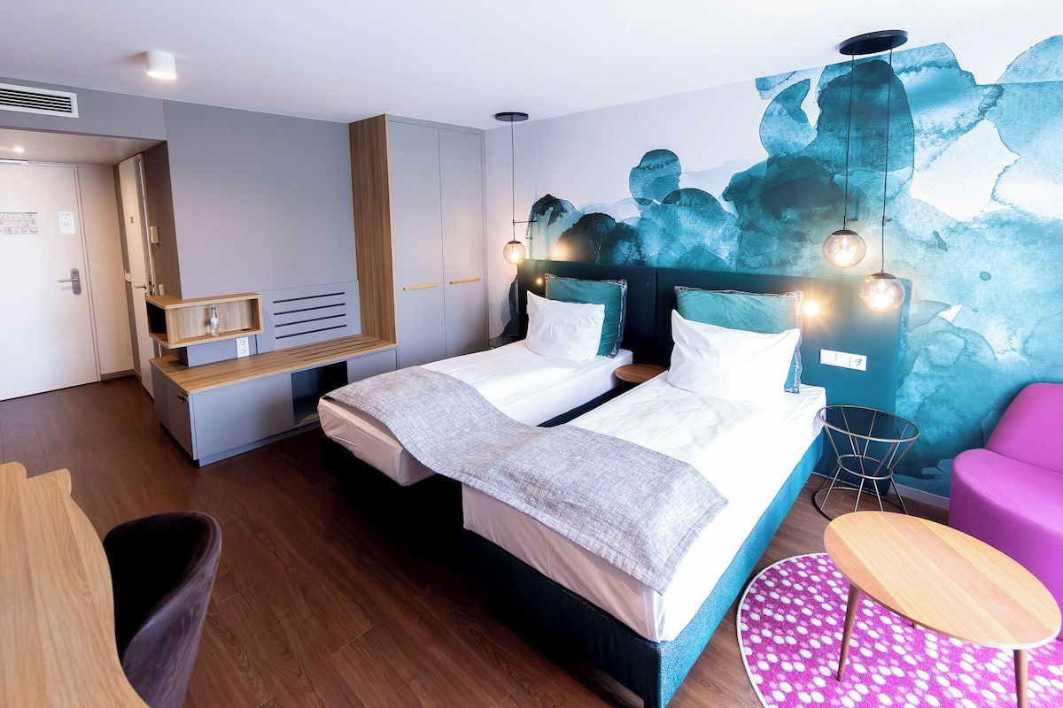 A modern hotel room with a bed, a desk, a chair, and a wall mural.