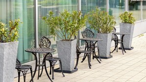Outdoor seating with tables and chairs, potted plants, and a building with large windows in the background. - DORMERO Hotel Freiburg (Freiburg im Breisgau)