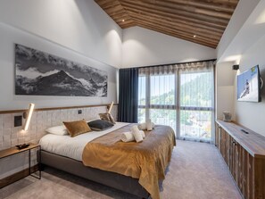 Quarto moderno com uma cama grande, uma pintura de montanhas, uma TV com tema de esqui e vista para uma paisagem montanhosa.