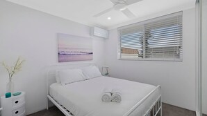 Ein Schlafzimmer mit Bett, Deckenventilator, Klimaanlage, Fenster mit Jalousien und einem Wandbild.