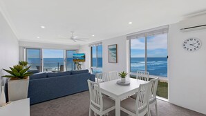 Ein modernes Wohnzimmer mit einer Couch, einem Esstisch und Stühlen, großen Fenstern mit Blick auf den Ozean und einem Deckenventilator.
