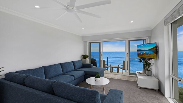 Ein modernes Wohnzimmer mit Ecksofa, Deckenventilator, Fernseher und Meerblick durch große Glasfenster.