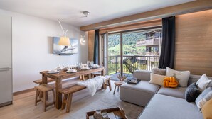 A modern living room with a wooden dining table set for four, a sofa with decorative pillows, and a large window offering a view of a mountain landscape. - Luxury 3-bedroom apartment in Megève with pool and gym (Megève)