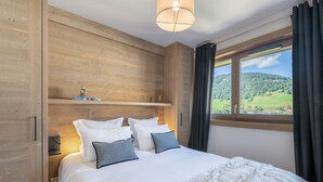A bedroom with a wooden headboard, a bed with white bedding and two pillows, a small shelf with decorative items, and a window with a view of a green landscape. - Luxury 3-bedroom apartment in Megève with pool and gym (Megève)