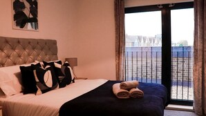 Un dormitorio con cabecera capitonada, cama con almohadas blancas y negras, colcha oscura y un par de toallas enrolladas.