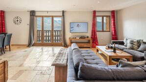 A spacious living room with a large sofa, a coffee table, and a television. There is a dining area with chairs and a table, and a view of mountains through the glass doors. - Spacious apartment 6 pers, terrace sauna close to slopes (Val Thorens)