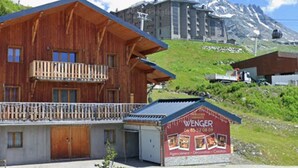 A wooden chalet with a balcony, a small attached building with a red sign, and a mountainous backdrop. - Spacious apartment 6 pers, terrace sauna close to slopes (Val Thorens)