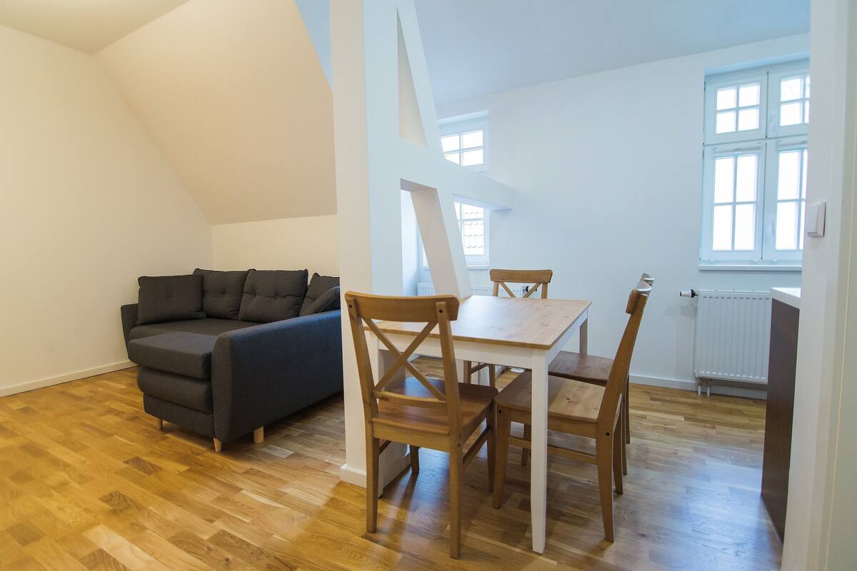 Ein Zimmer mit Holzboden, einem dunkelgrauen Sofa, einem Holzesstisch mit vier Stühlen und einem Fenster mit weißen Fensterläden.