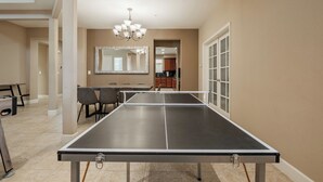 A ping pong table in a room with a dining area, a chandelier, and a kitchen in the background. - Tesla EV Charger Solar heated Pool & Hot Tub Near UC Davis, Med Center, Airport (West Sacramento)