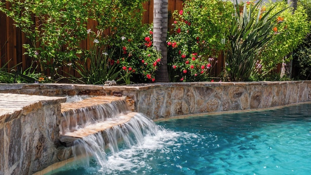 A swimming pool with a waterfall feature, surrounded by lush greenery and a wooden fence. - Tesla EV Charger Solar heated Pool & Hot Tub Near UC Davis, Med Center, Airport (West Sacramento)