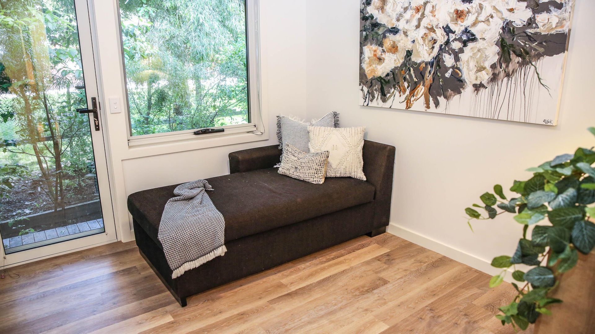 A cozy room with a dark brown sofa, a large window with a view of greenery, and a painting on the wall.