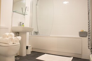 A modern bathroom with a white sink, toilet, and bathtub. There are towels stacked on the toilet, a glass shelf above the sink, and a small trash bin beside the toilet.
