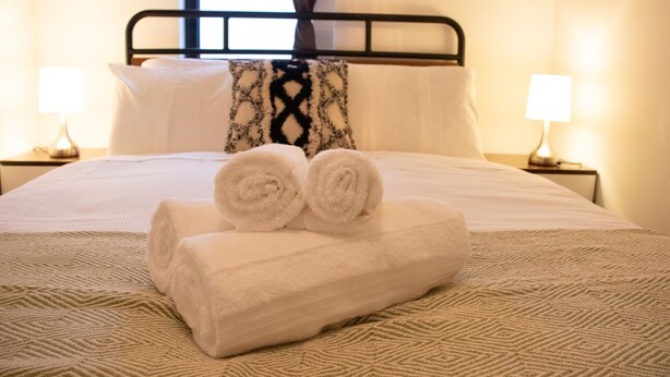 Cama bem arrumada com toalhas enroladas, colcha estampada e duas luminárias em mesas de cabeceira.
