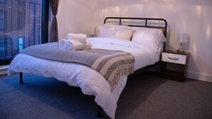 Cama bem arrumada com lençóis brancos e manta estampada, mesa de cabeceira com abajur, e janela com cortinas.