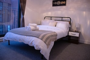 A neatly made bed with white linens and a patterned throw, a bedside table with a lamp, and a window with curtains.