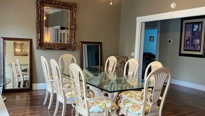 A dining room with a glass-top table surrounded by white chairs with floral upholstery, wooden flooring, and a ceiling fan.