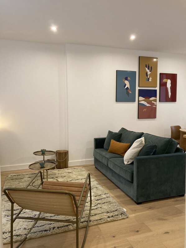 Un salon moderne avec un canapé vert foncé, une petite table basse ronde et une chaise en métal. Trois œuvres d’art encadrées ornent le mur, ainsi qu’une petite table d’appoint en bois et un tapis moelleux sur le sol.