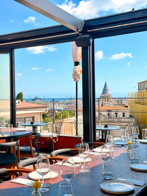 Restaurante com mesas preparadas para refeições, grandes janelas com vista para a cidade e céu claro.