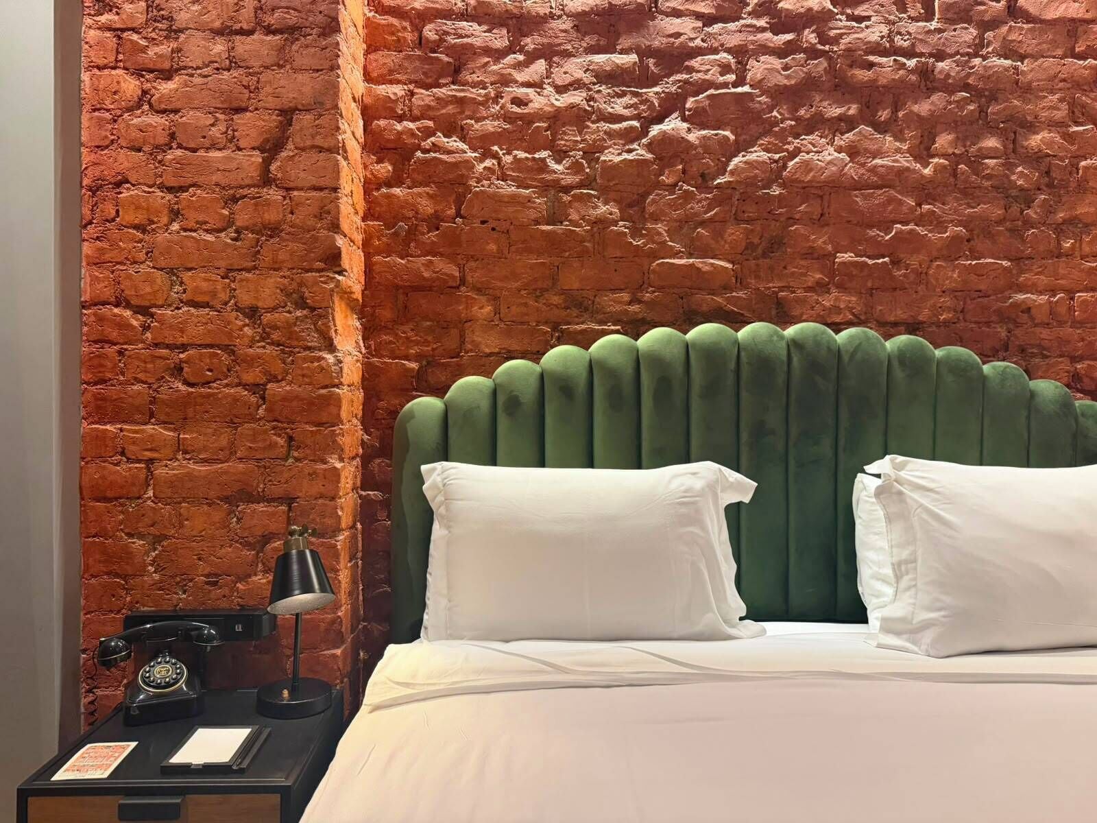 A room with a brick wall, a green upholstered headboard, and a classic telephone on a side table.