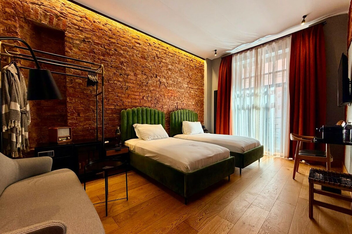 A hotel room with a brick wall, two beds, a wooden floor, a TV, and a desk with a chair.